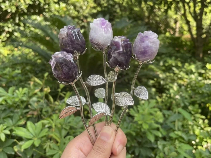 Hands supporting five purple and lavender crystal rosebuds on silver-colored stems and a small flared stand against a blurred green background