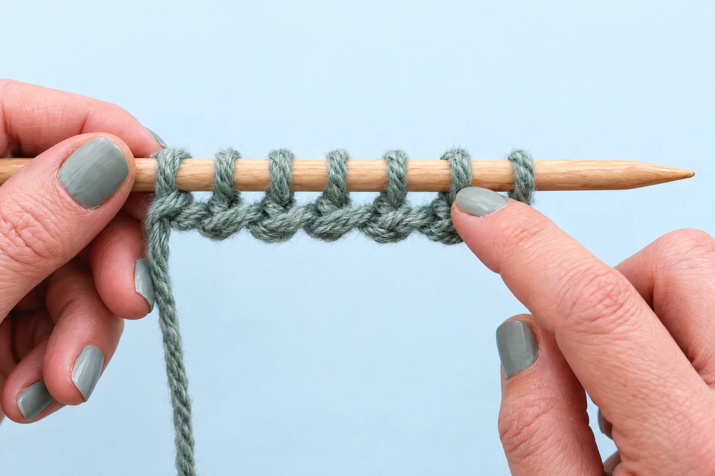 Hands gently check a short sample of spaced sage-green cast-on loops along the full shaft of a wooden needle.