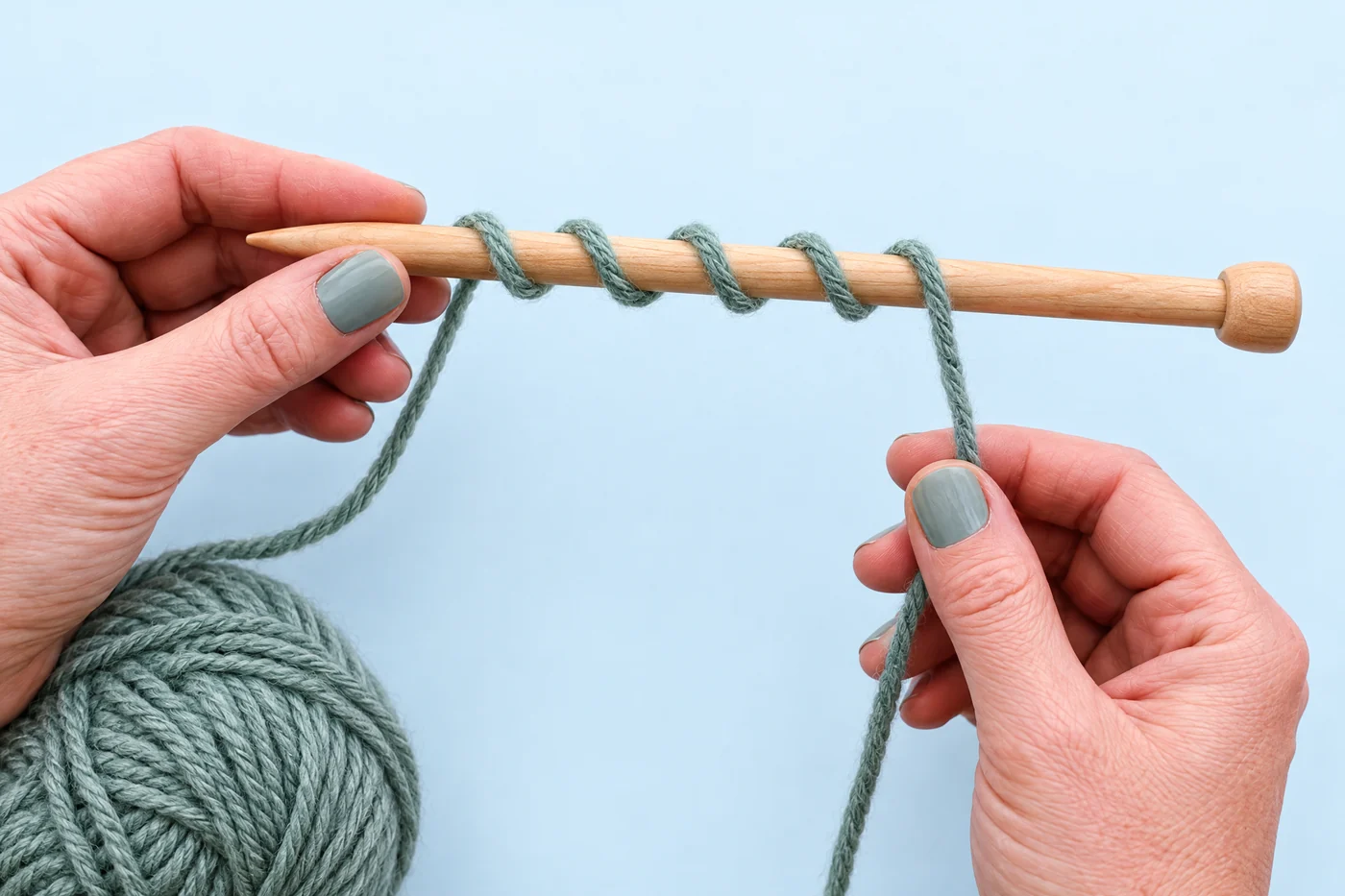 Several loose sage-green yarn wraps surround a wooden knitting needle held above a matching yarn ball.