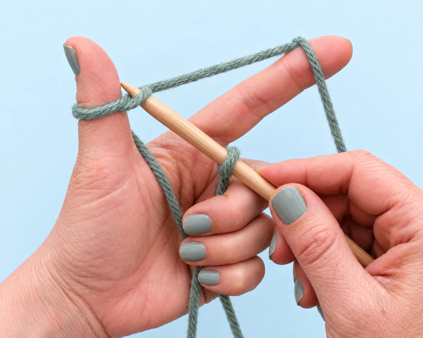 A pointed wooden knitting needle lifts the near strand of a sage-green thumb loop from below during long-tail cast-on.